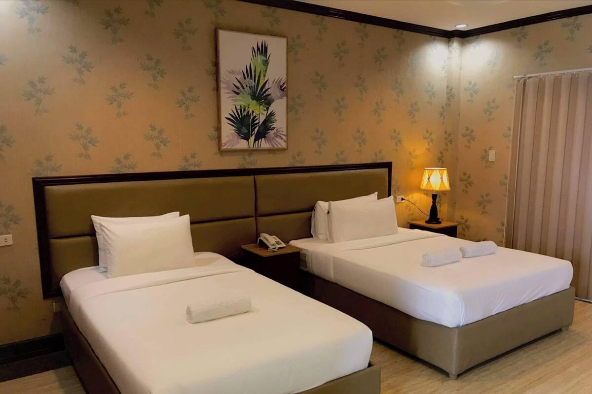 Una habitación de hotel con dos camas, una mesita de noche con lámpara y un papel tapiz floral.