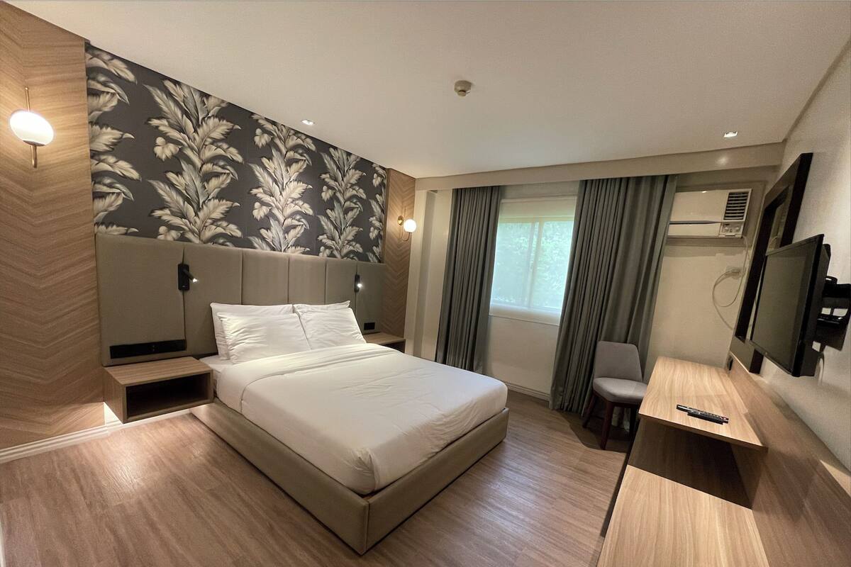 Una habitación de hotel moderna con una cama grande, una mesita de noche, una silla, un televisor y una ventana con cortinas.
