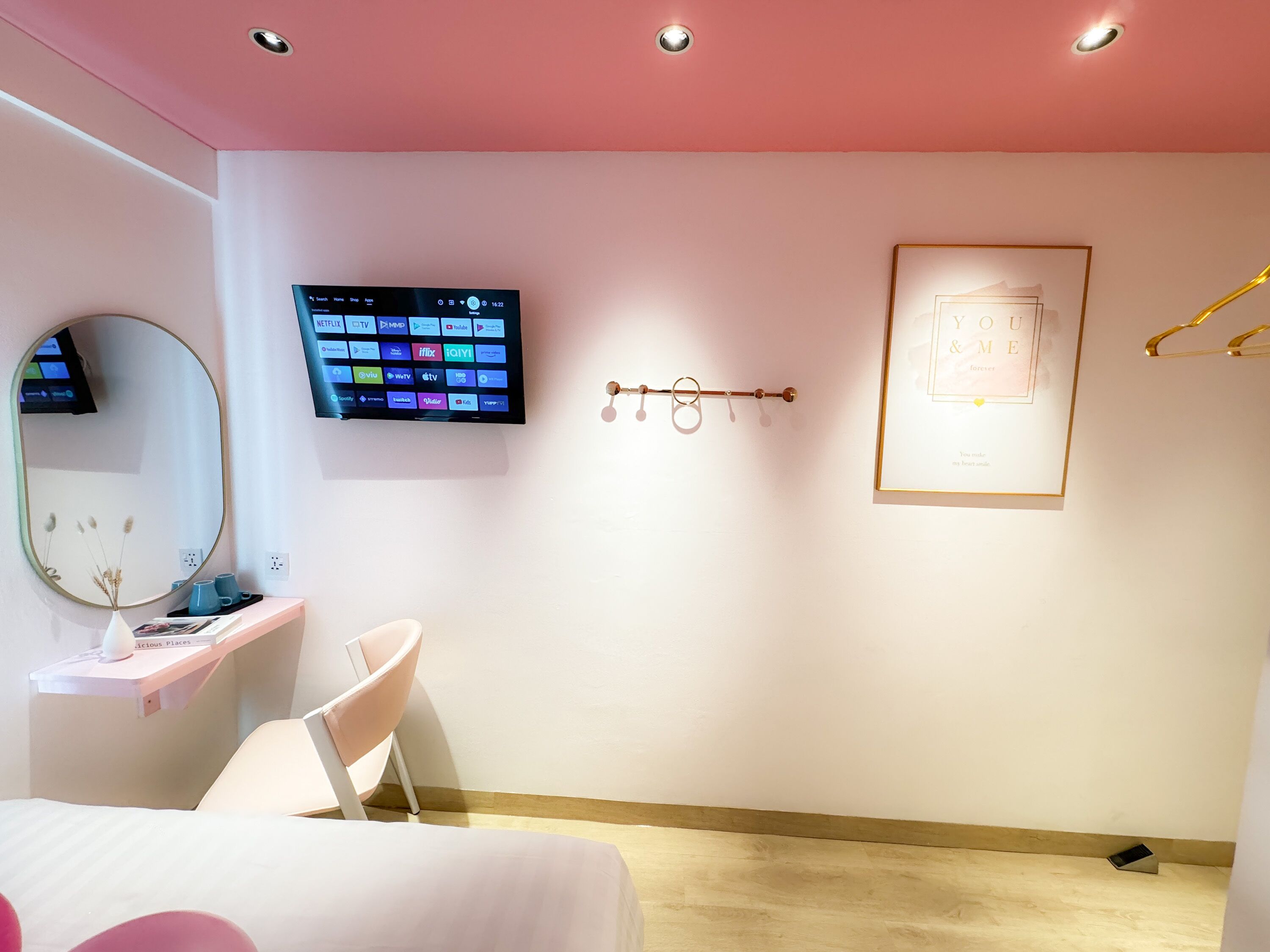 Habitación de hotel moderna con techo rosa, televisor de pantalla plana en la pared, mesita de noche con un jarrón y libros, una silla blanca y una cita enmarcada en la pared.