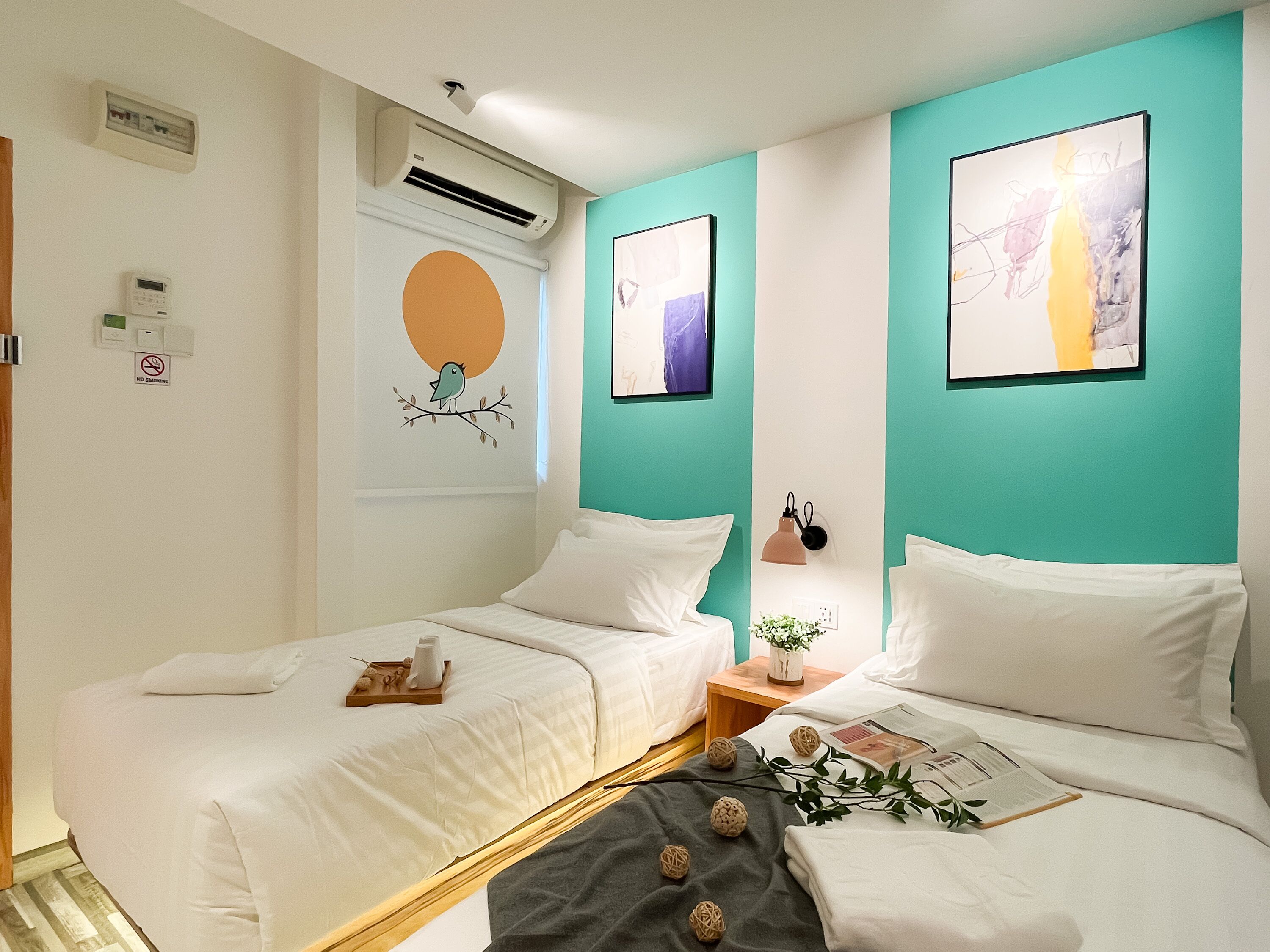 A hotel room with two beds, a wall-mounted air conditioner, a no-smoking sign, and artwork.