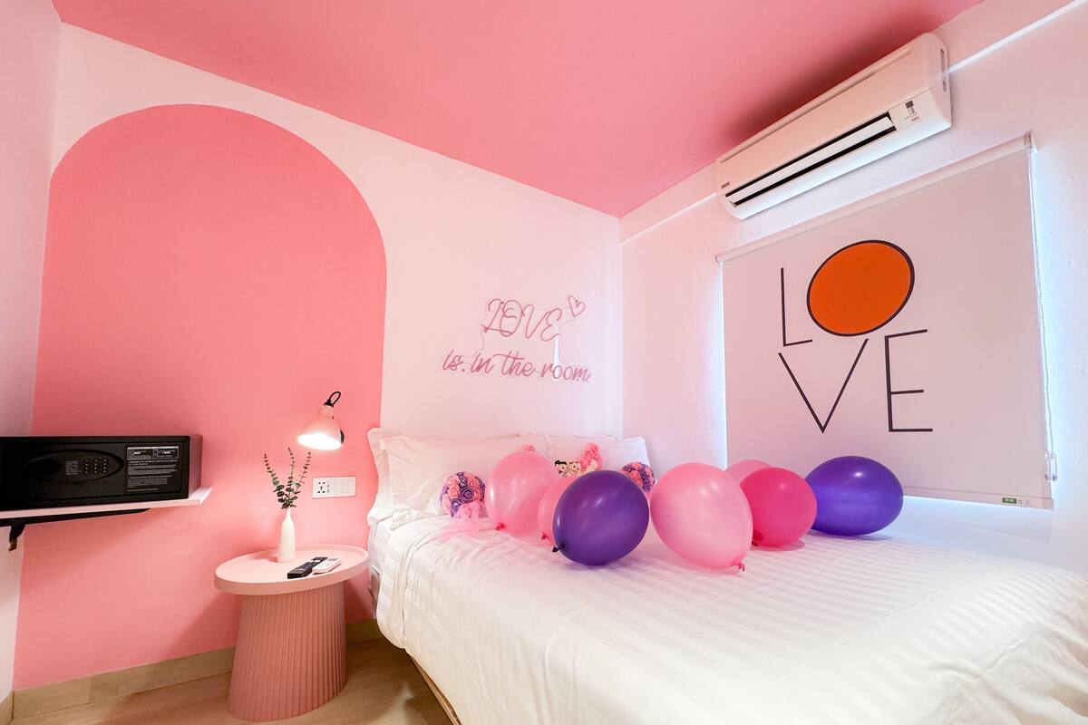 A pink-themed bedroom with a bed, pillows, balloons, a bedside table with a lamp, and a wall-mounted air conditioner.