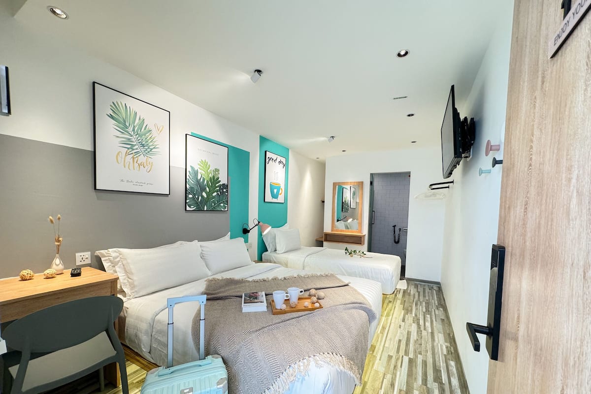 A hotel room with two beds, a desk, a chair, a suitcase, and a bathroom with a mirror and a shower.