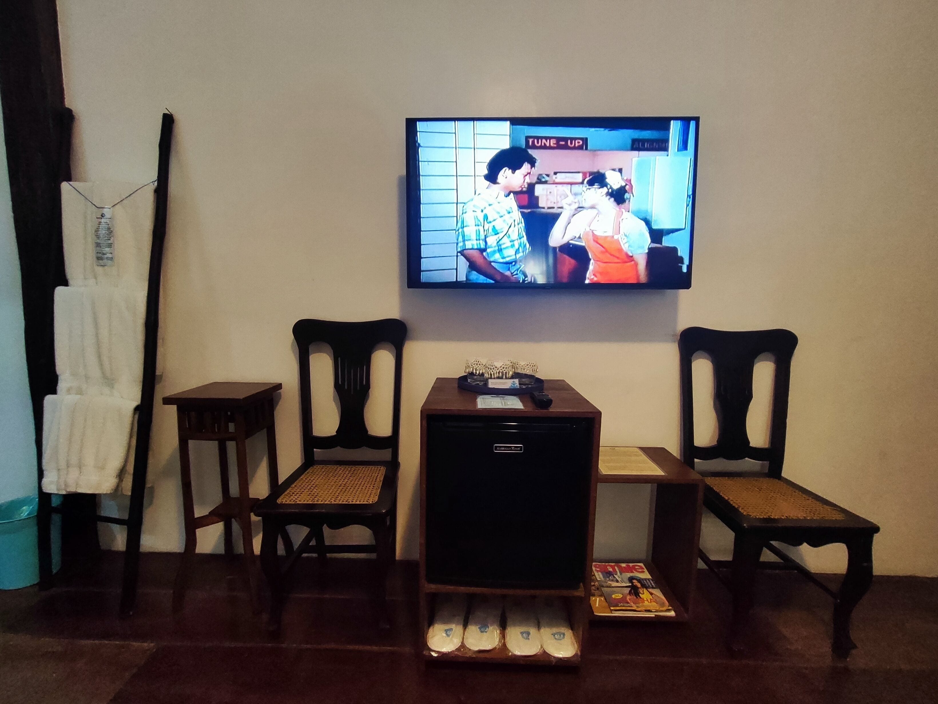 Una sala de estar con una televisión de pantalla plana montada en la pared, dos sillas de madera, una mesita de madera y una estantería con revistas y un control remoto.