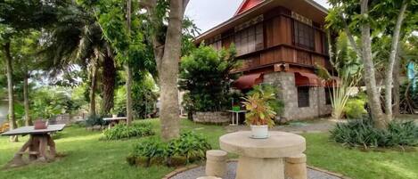 A traditional wooden house surrounded by lush greenery and a circular stone seating area with a central table.