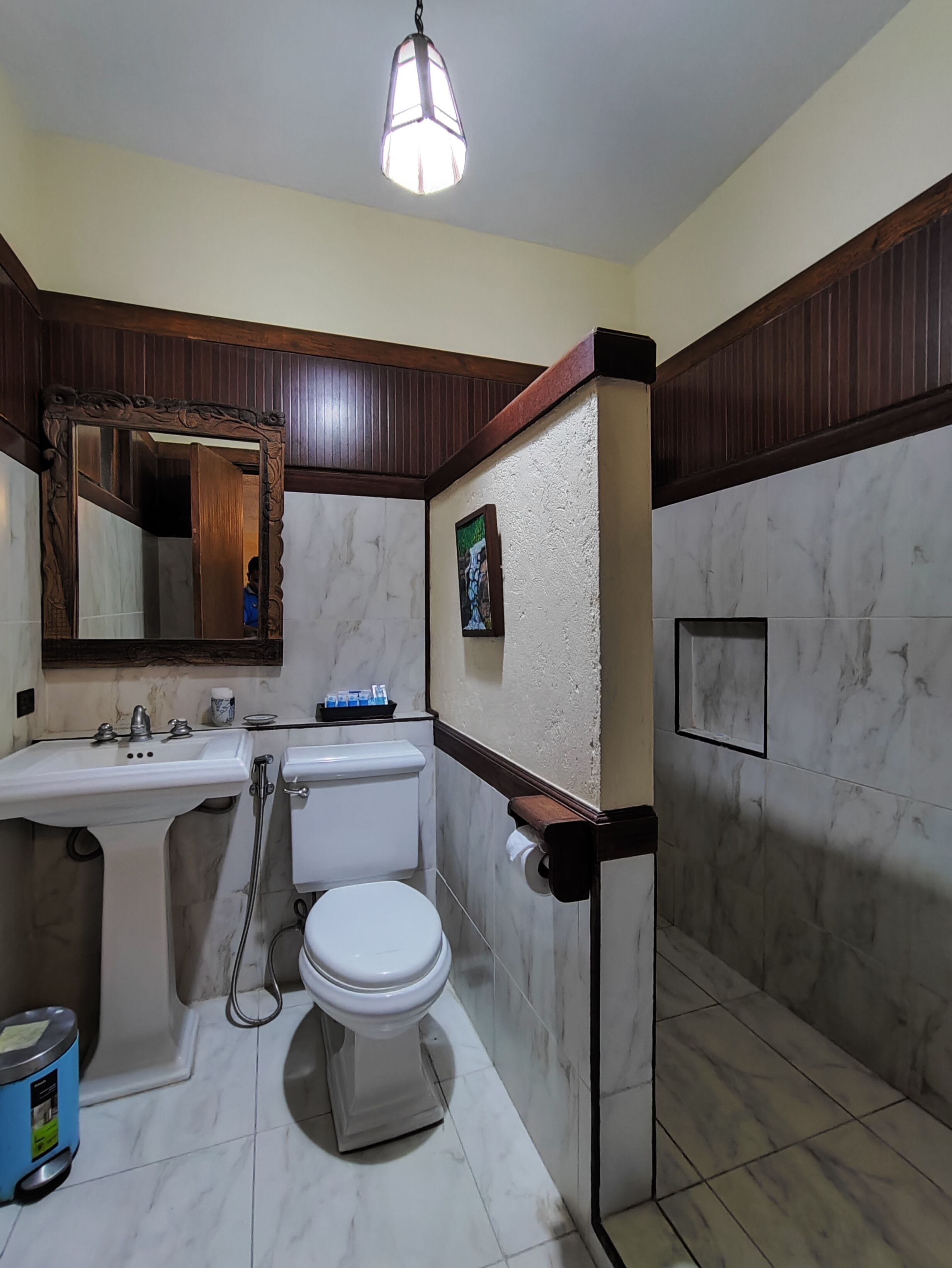 A bathroom with a white sink, toilet, and shower. There is a mirror above the sink, a wooden-framed mirror, and a picture on the wall.