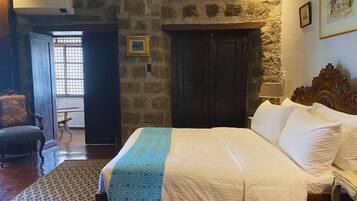 A bedroom with a bed, a chair, a wooden wardrobe, and a stone wall.