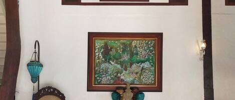 Une pièce agrémentée d’un lustre, d’un tableau botanique encadré, d’un meuble en bois orné d’objets décoratifs, d’une chaise en osier et d’un vase bleu.