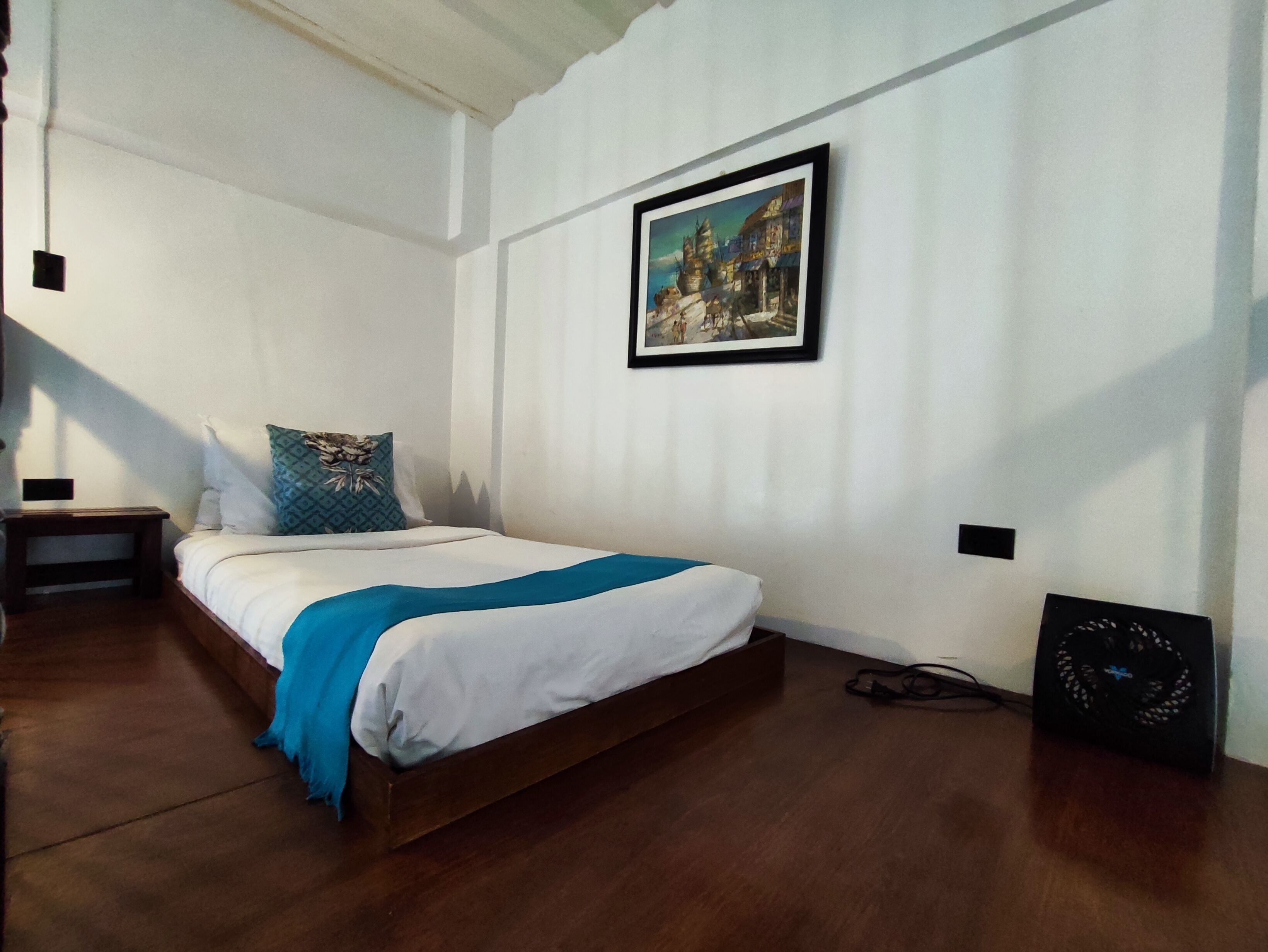 A bedroom with a wooden bed, white walls, a framed painting, and a small table with a lamp.