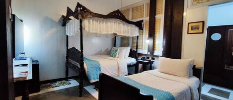A traditional four-poster bed with a canopy, a desk with a lamp, and a patterned floor.