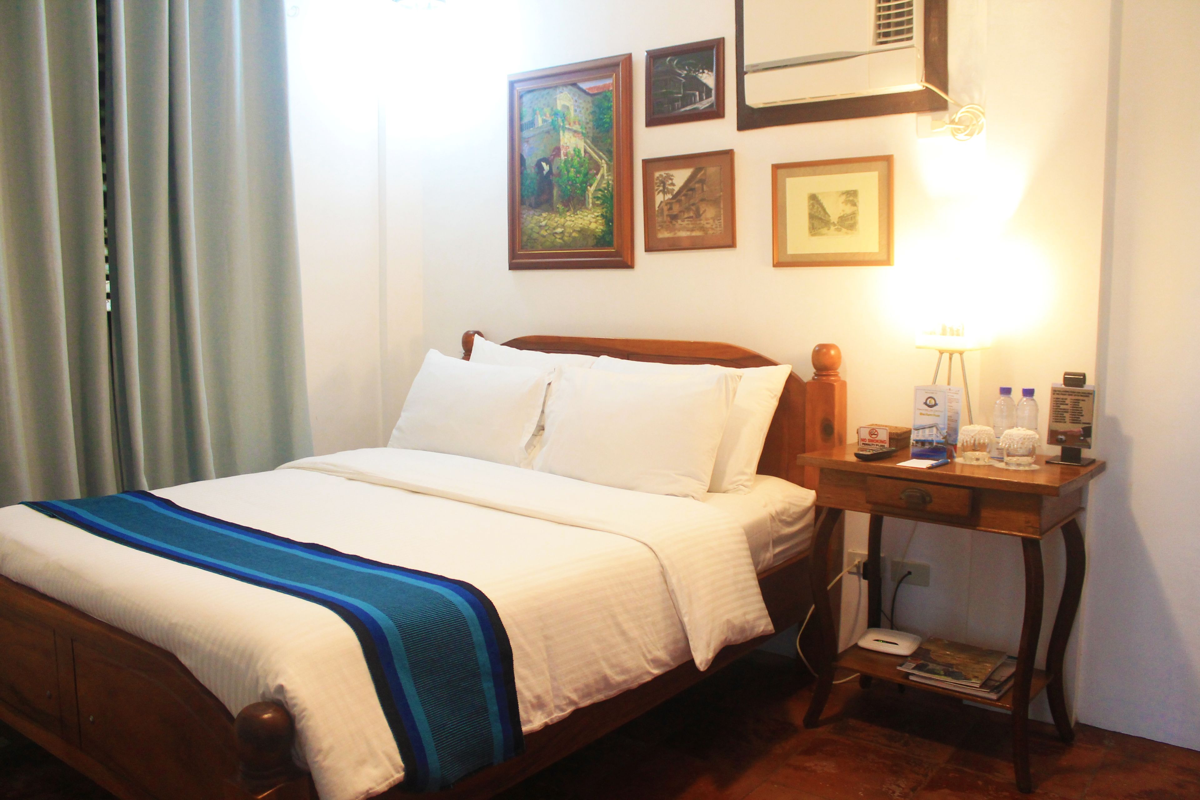 Een net opgemaakt bed met een blauw-wit gestreept dekbed, een houten nachtkastje met een lamp, en ingelijste foto's aan de muur.