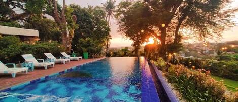 A swimming pool with blue tiles, surrounded by greenery and trees, with a sunset in the background.