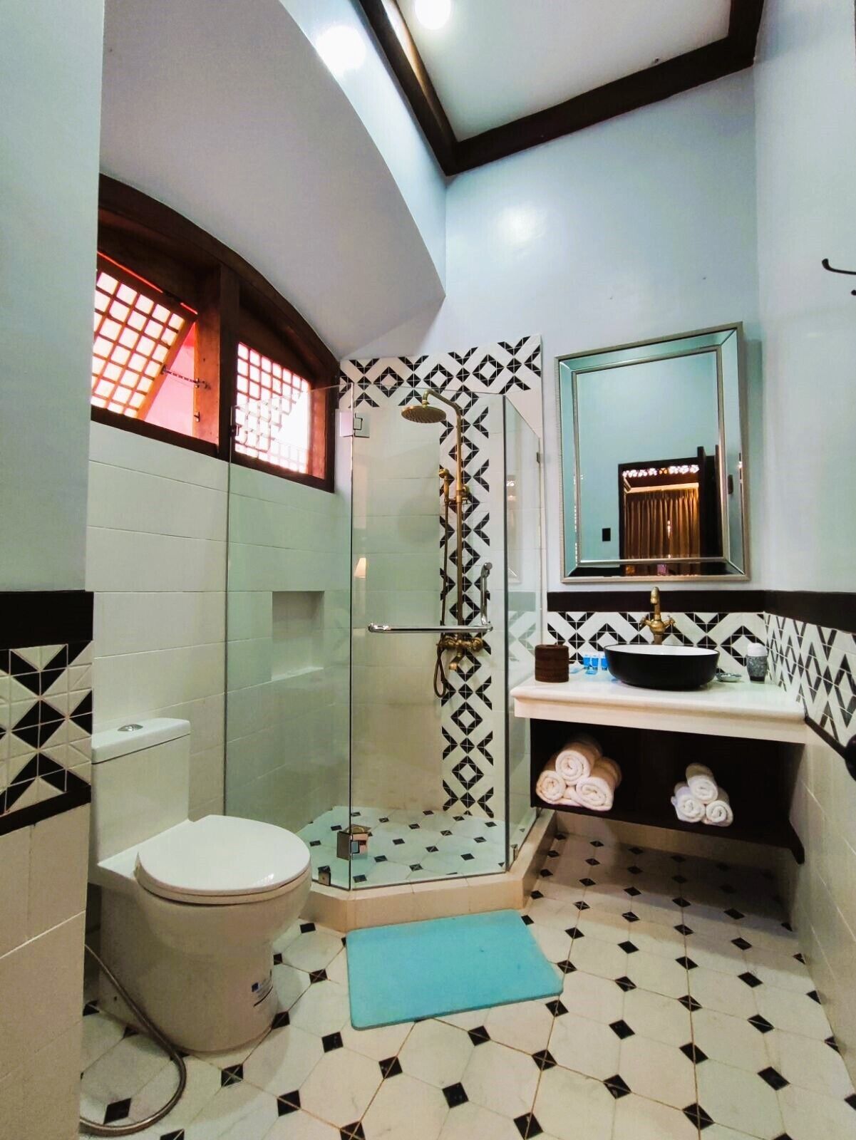 Un baño con ducha envuelta en vidrio, inodoro, lavamanos con espejo sobre él y piso estampado.