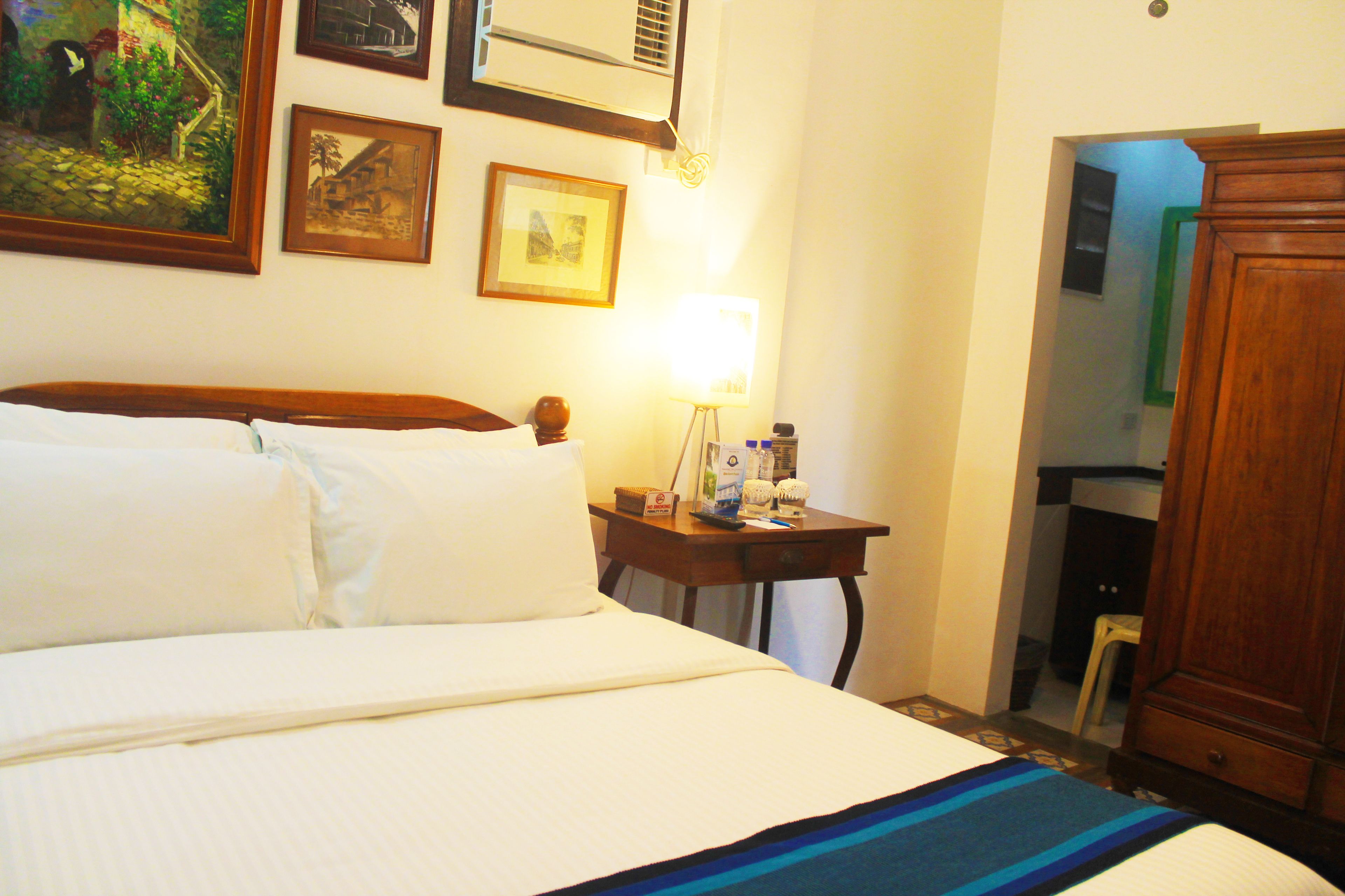 Una camera d'albergo con un letto, un comodino con una lampada, un armadio in legno e quadri appesi al muro.