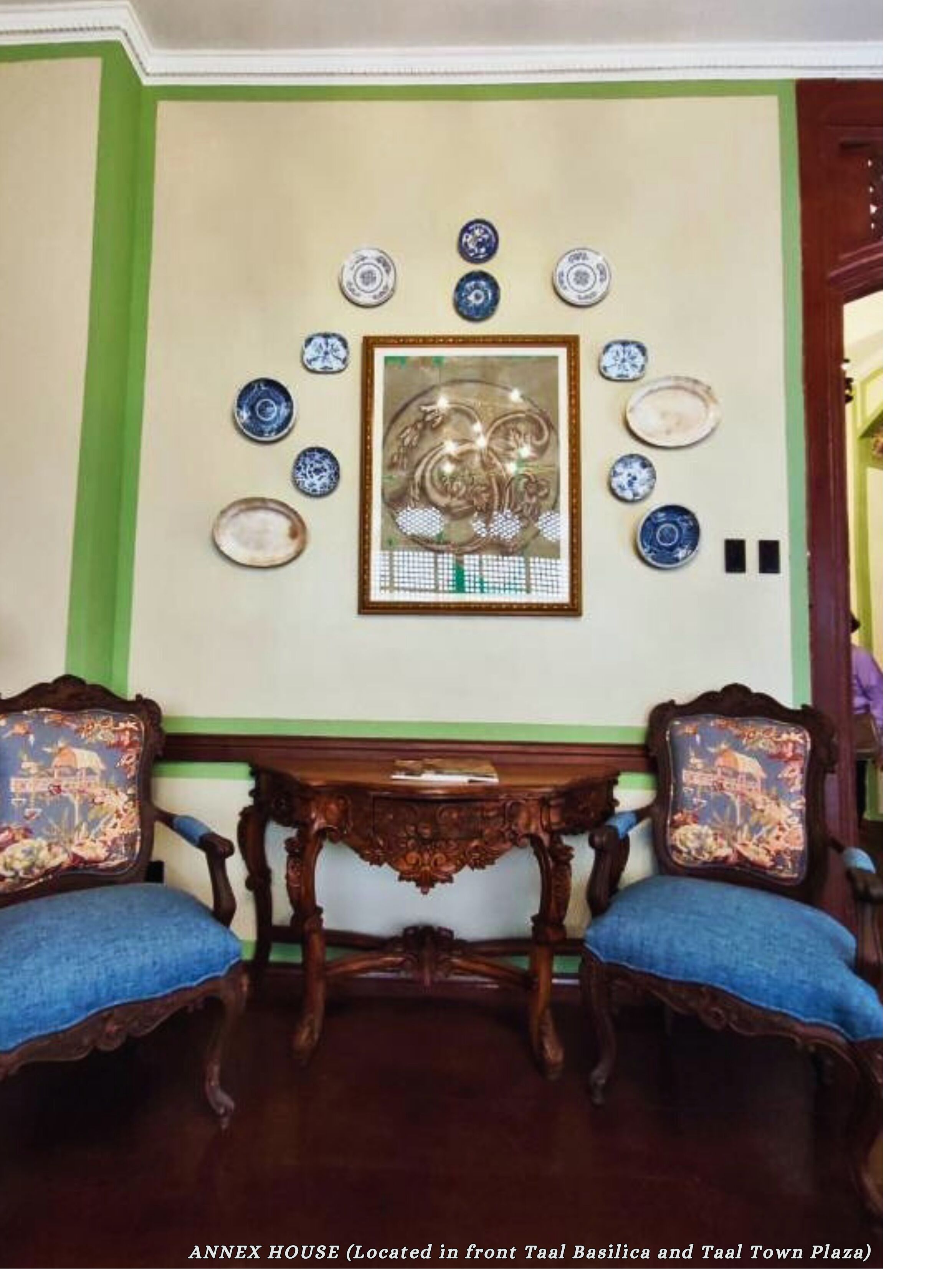 Um quarto com duas cadeiras ornamentadas, uma mesa de madeira e uma parede decorada com pratos e uma obra de arte emoldurada.