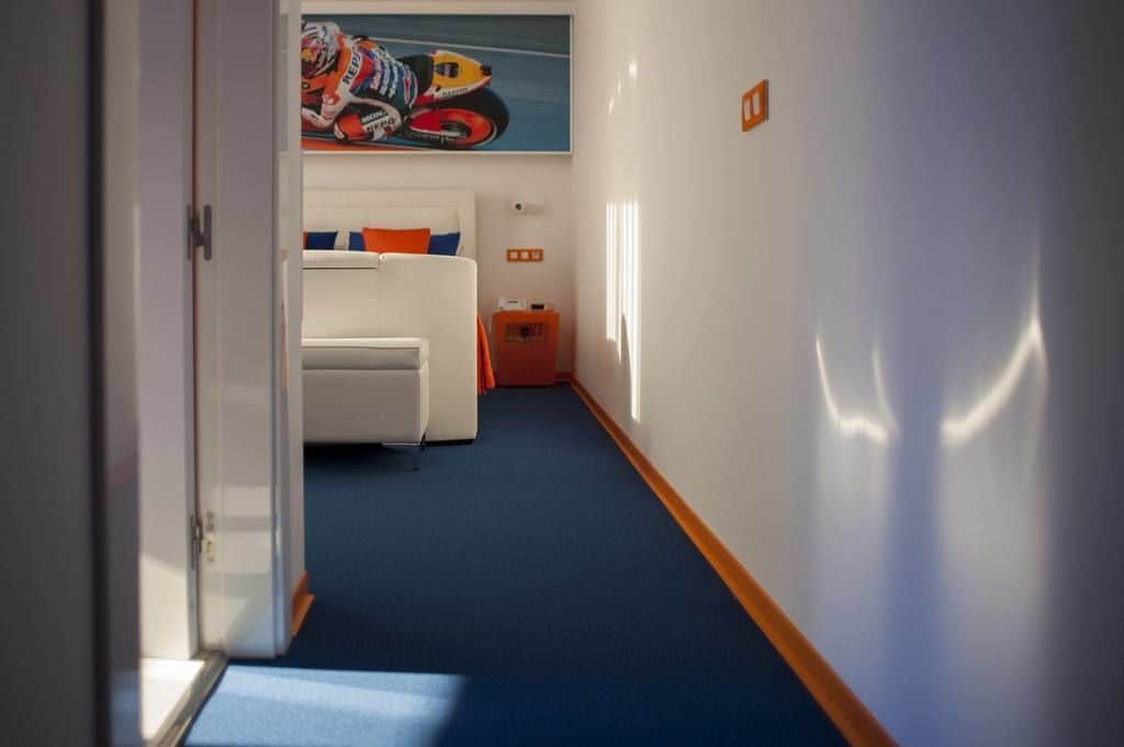 A hotel corridor with a blue carpet, a white bed with orange pillows, and a motorcycle racing poster on the wall.