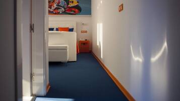 A hotel corridor with a blue carpet, a white bed with orange pillows, and a motorcycle racing poster on the wall.