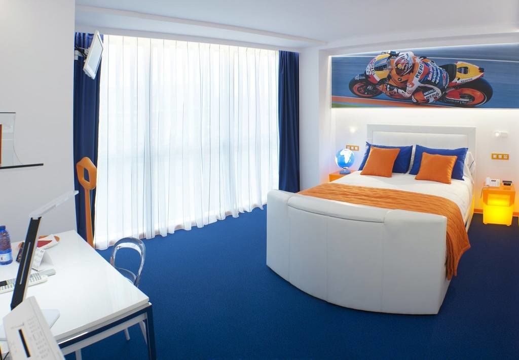 A modern hotel room with a large bed, a desk with a chair, a globe, and a motorcycle-themed artwork on the wall.