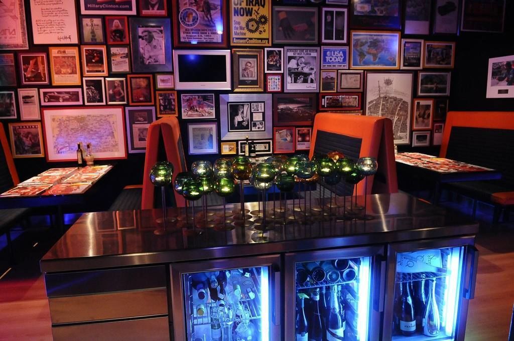 Uma área de bar com uma parede de fotografias emolduradas, duas cadeiras laranja e uma mesa com uma garrafa e copos.