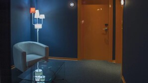 Habitación de hotel con una silla moderna, una mesa de centro de vidrio y una puerta con interruptor de luz.