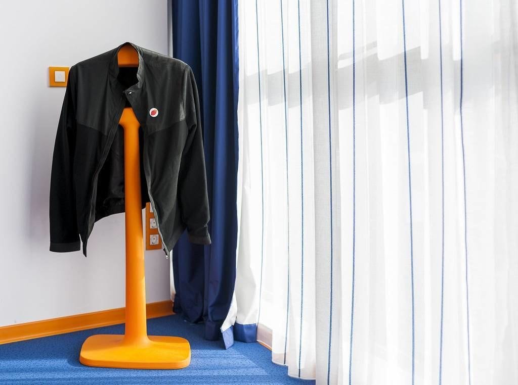 Une pièce avec une moquette bleue, une veste noire posée sur un porte-vêtements et une fenêtre ornée de rideaux blancs.