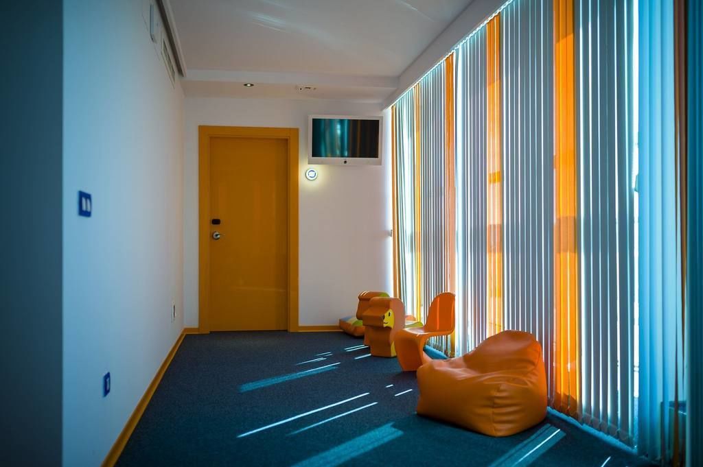 Een gang met een blauw tapijt, een houten deur en oranje zitplaatsen bij een raam met verticale jaloezieën.