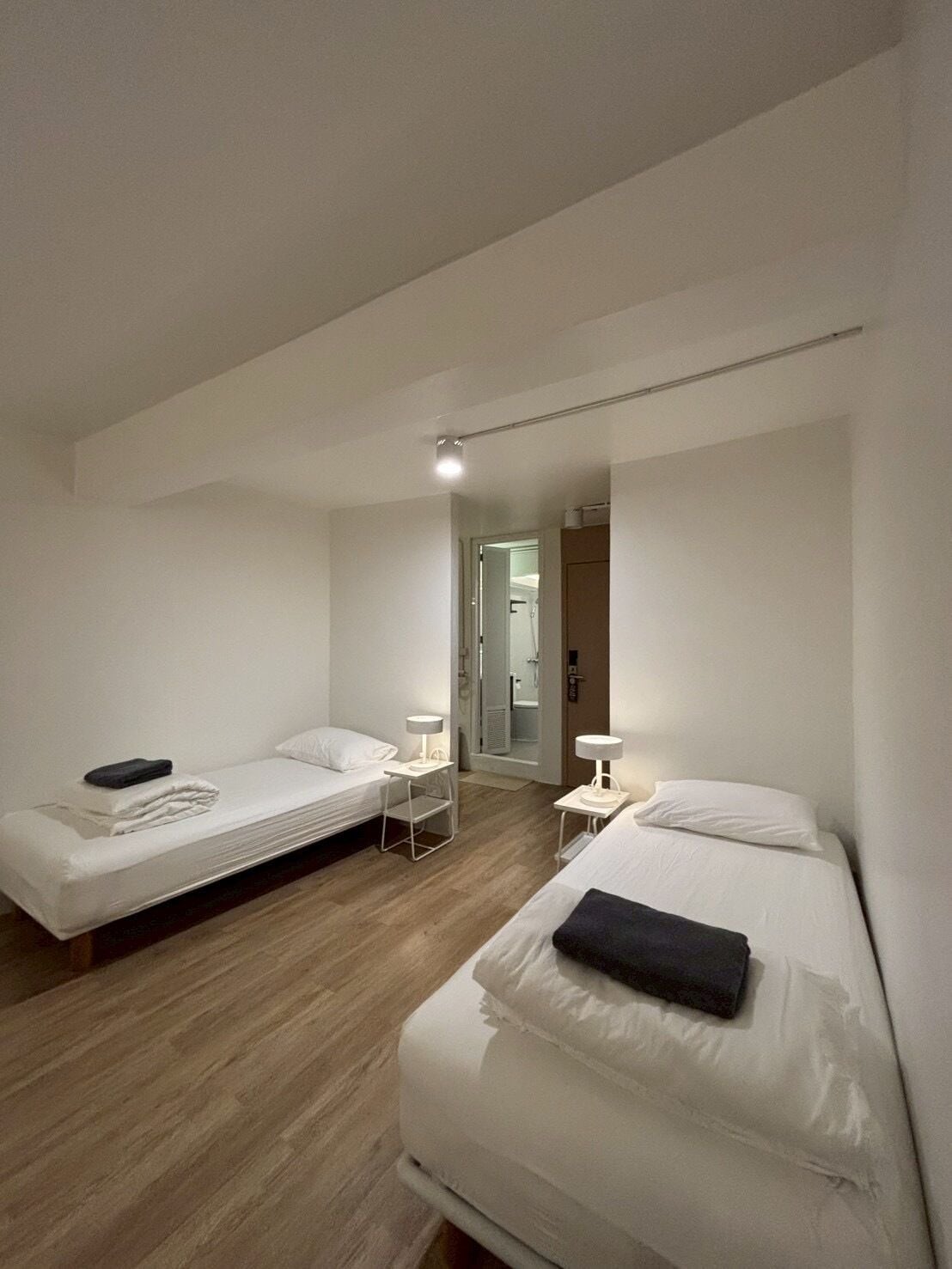 A minimalist hotel room with two single beds, a small table, a chair, and a bathroom visible through an open door.