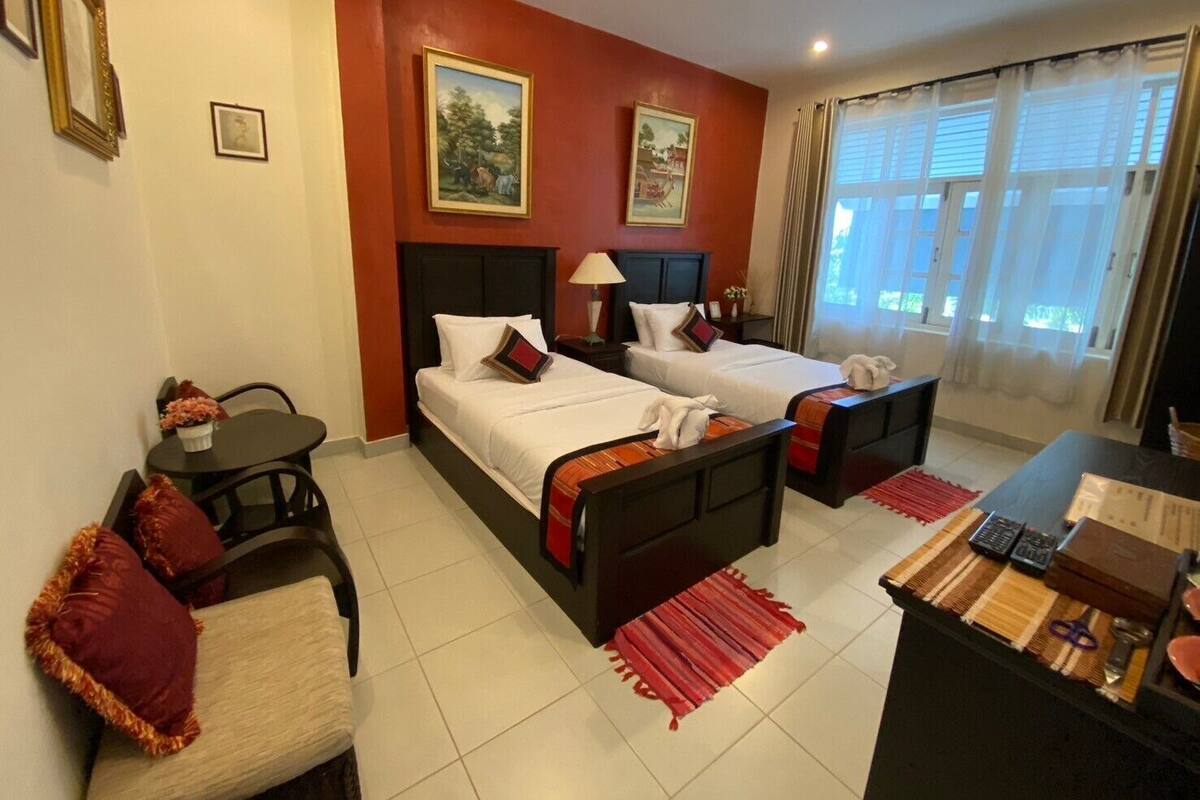 Une chambre d’hôtel avec deux lits, un bureau, une chaise et une petite table. Il y a deux tableaux au mur et une fenêtre avec des rideaux.