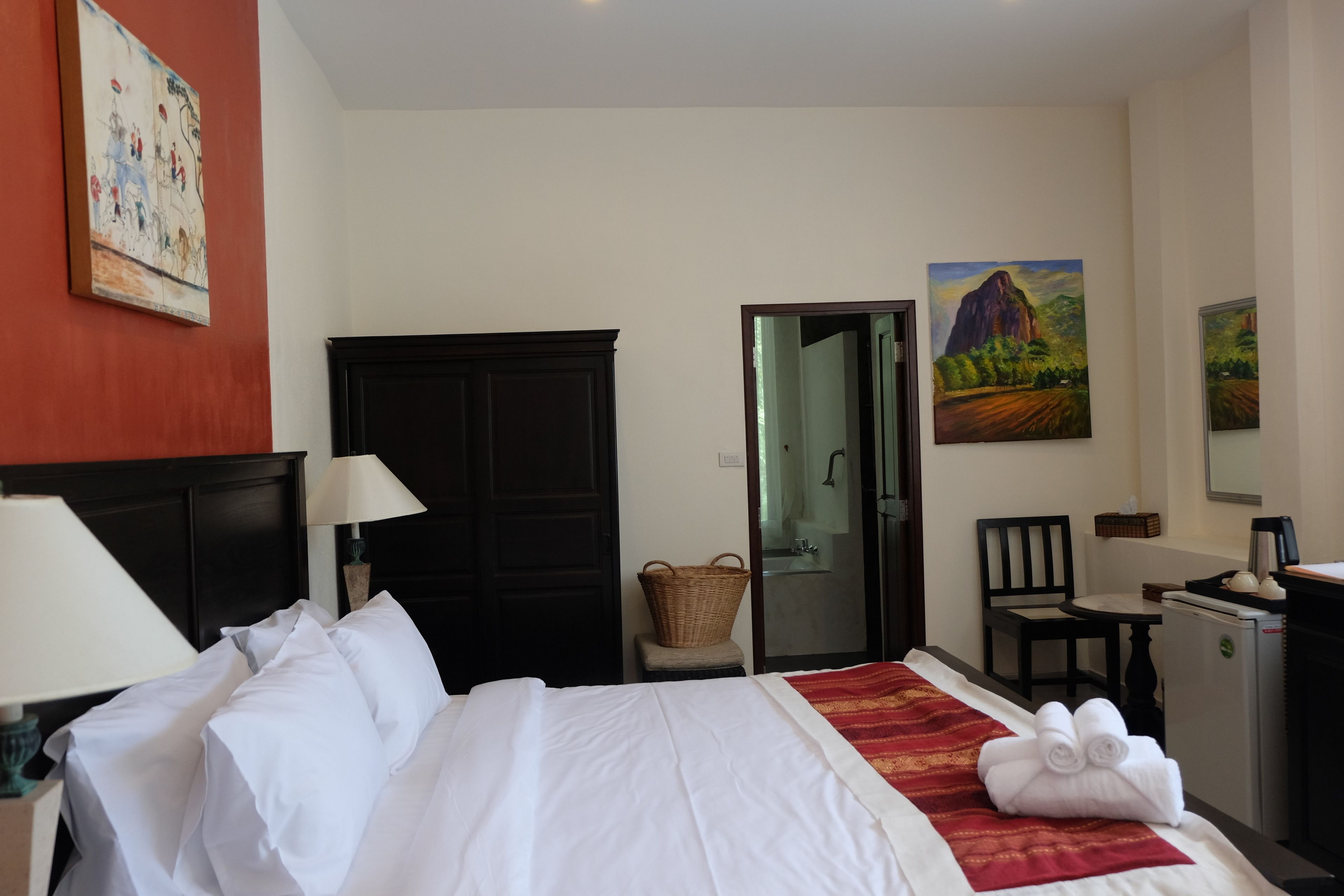 A hotel room with a bed, a desk, a chair, a lamp, a wardrobe, and two paintings on the wall.
