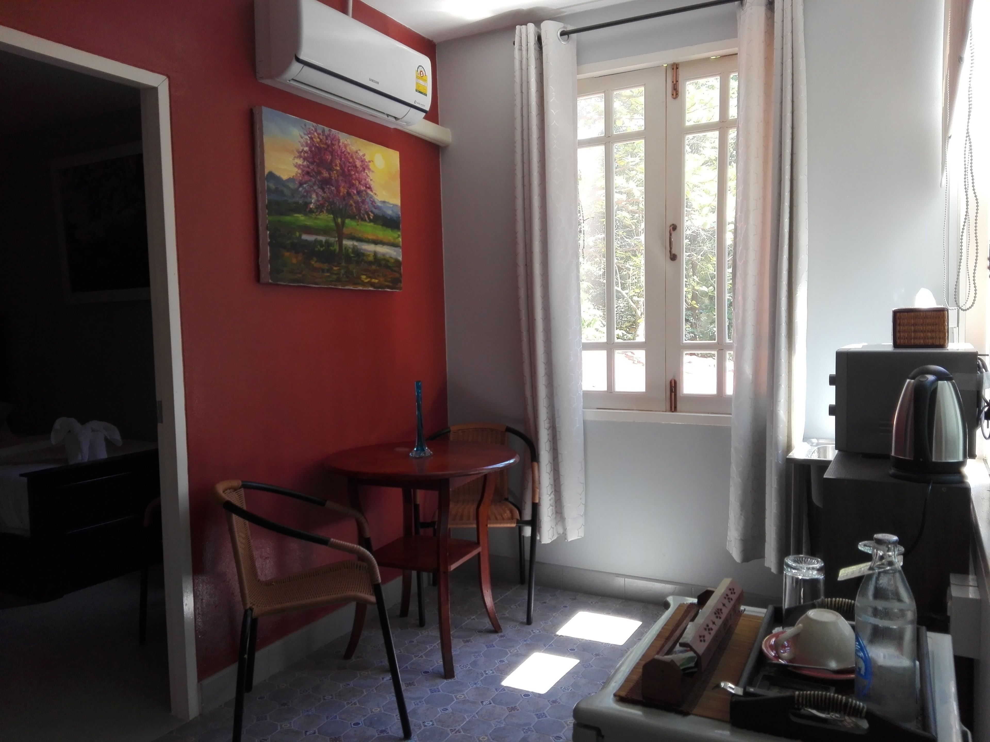 A small room with a red wall, a painting, a window with curtains, a table with a kettle, and a TV.