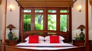 Una cama grande con cabecera de madera, dos almohadas rojas y vistas al verdor a través de amplias ventanas.
