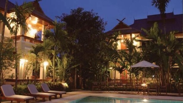 Zona de piscina con sillones de descanso y sombrillas, rodeada de palmeras y edificios con iluminación cálida.