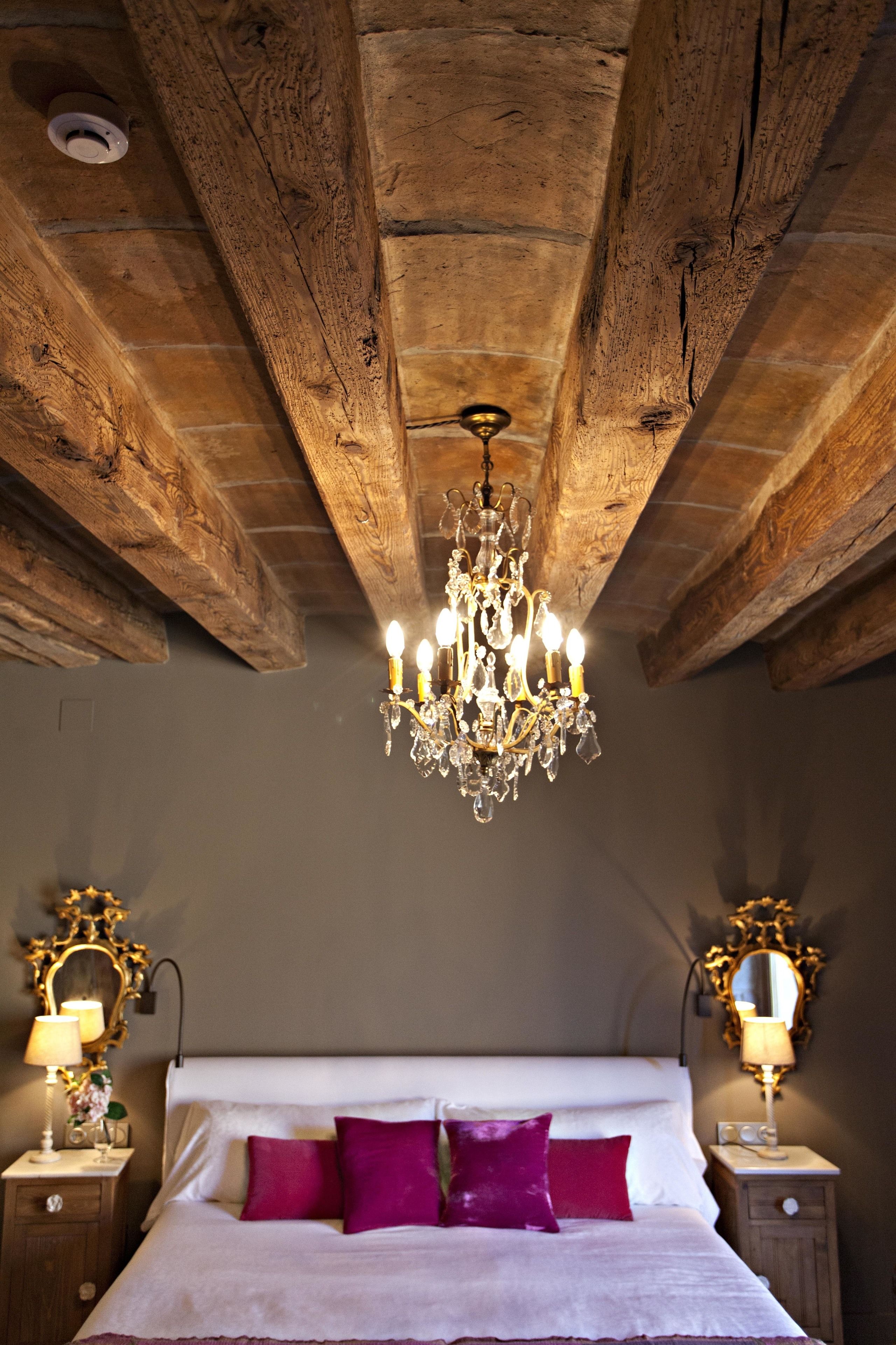 Un dormitorio con techo de madera, un candelabro, una cama con ropa de cama blanca y rosa, y mesitas de noche con lámparas.