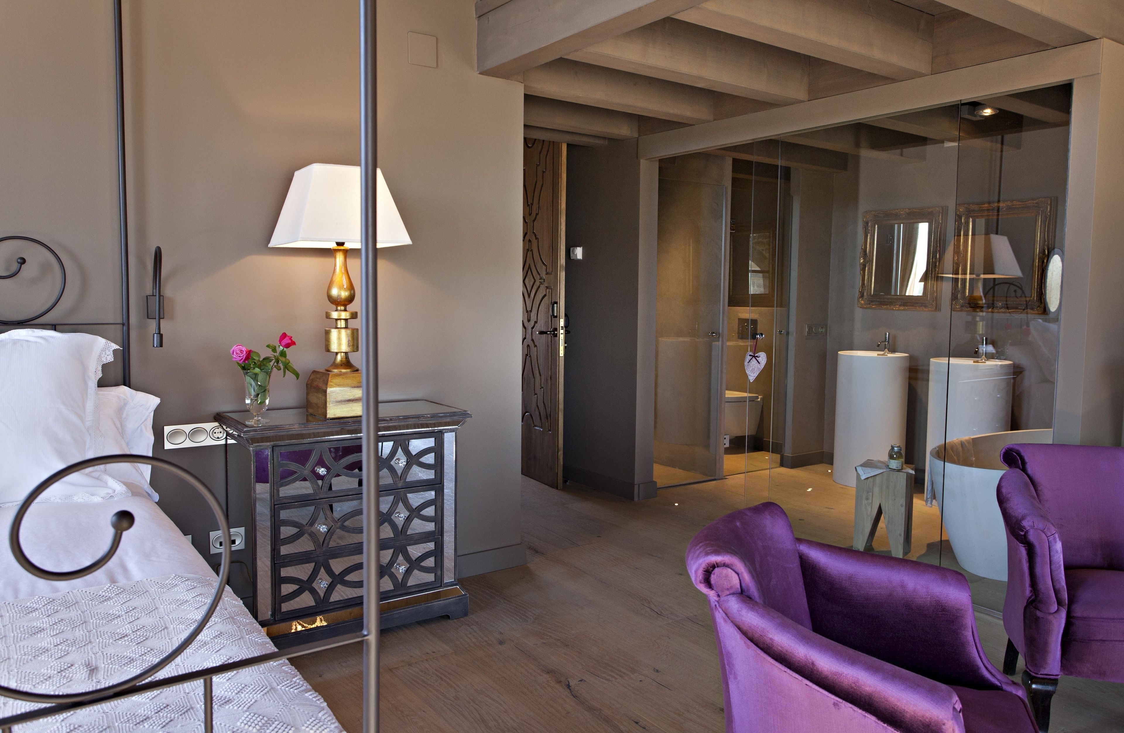 Una habitación de hotel moderna con una cama, una mesita de noche con lámpara, un sillón morado y un baño visible a través de una puerta de cristal.