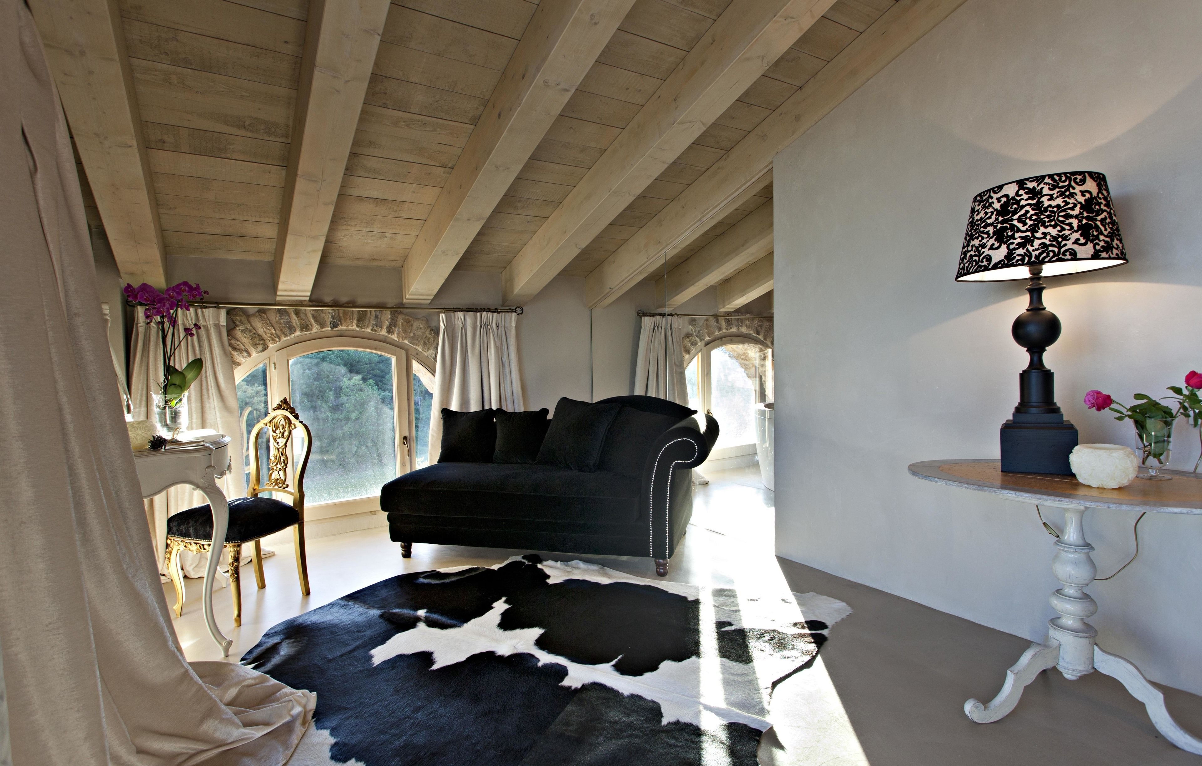 A room with a black sofa, a cowhide rug, a white table with a lamp, and a chair with a vase of flowers.