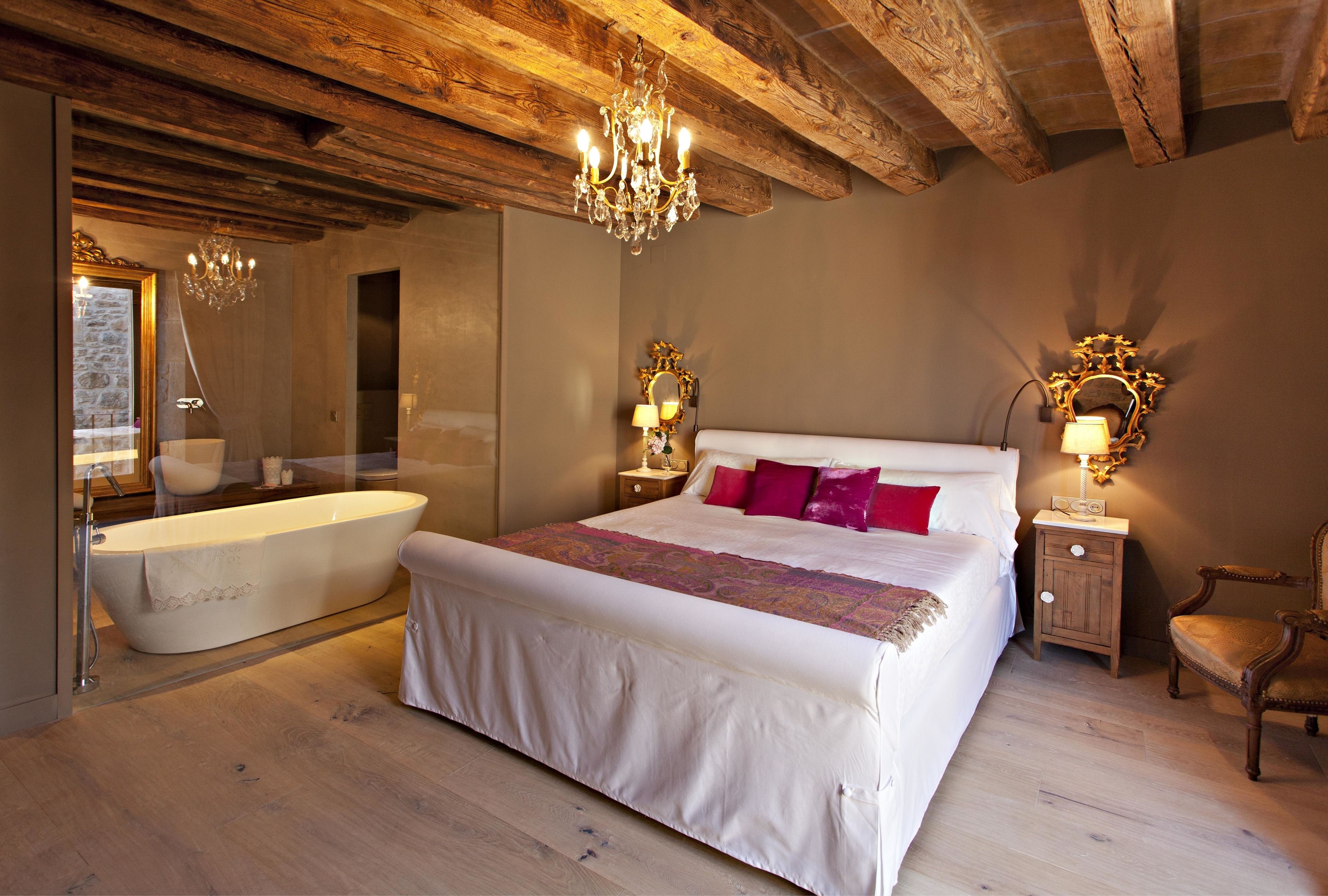 A bedroom with a large bed, a bathtub, wooden beams, and chandeliers.