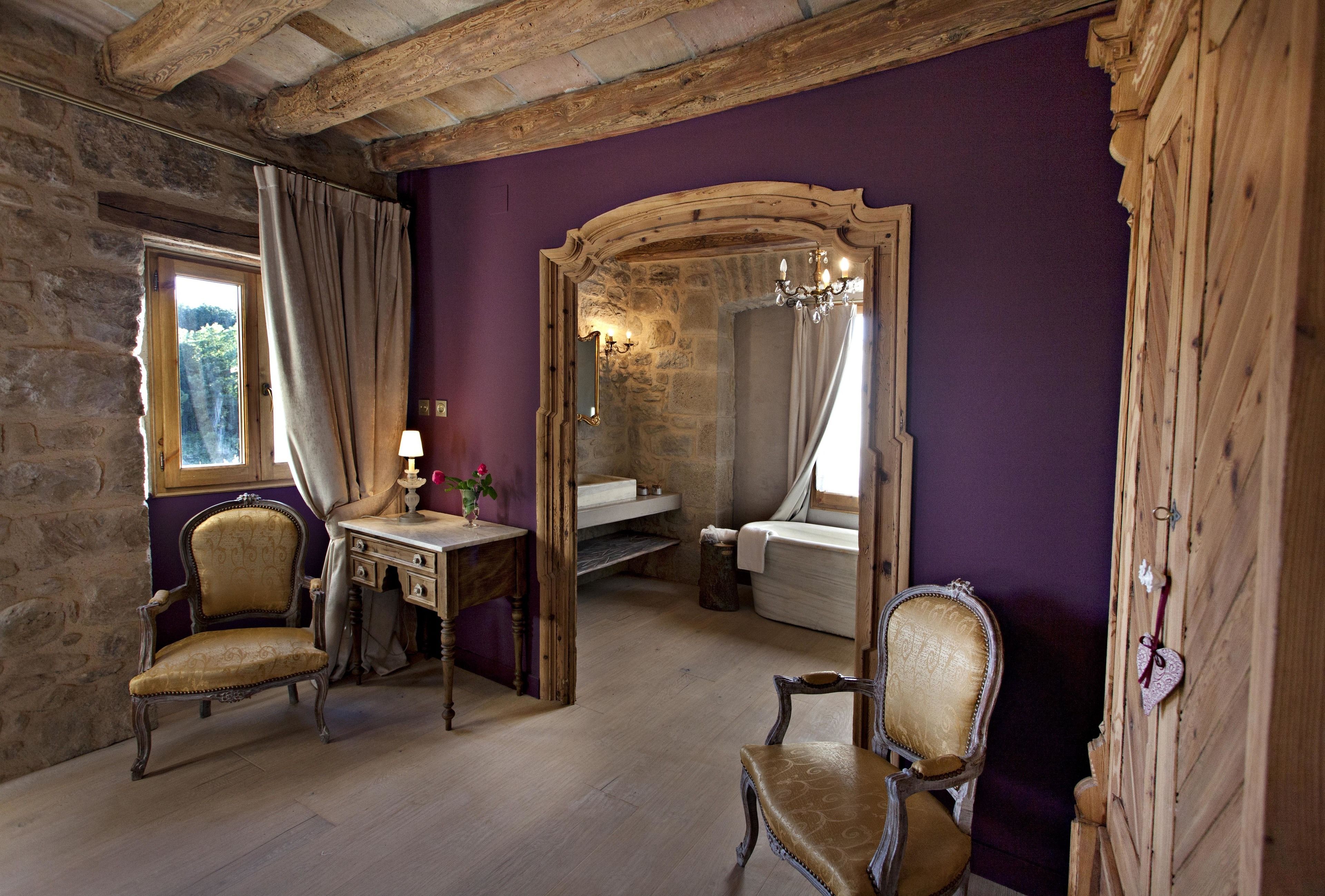 Una habitación con paredes de piedra, techo de madera, pared morada, una cama, un escritorio y dos sillas.