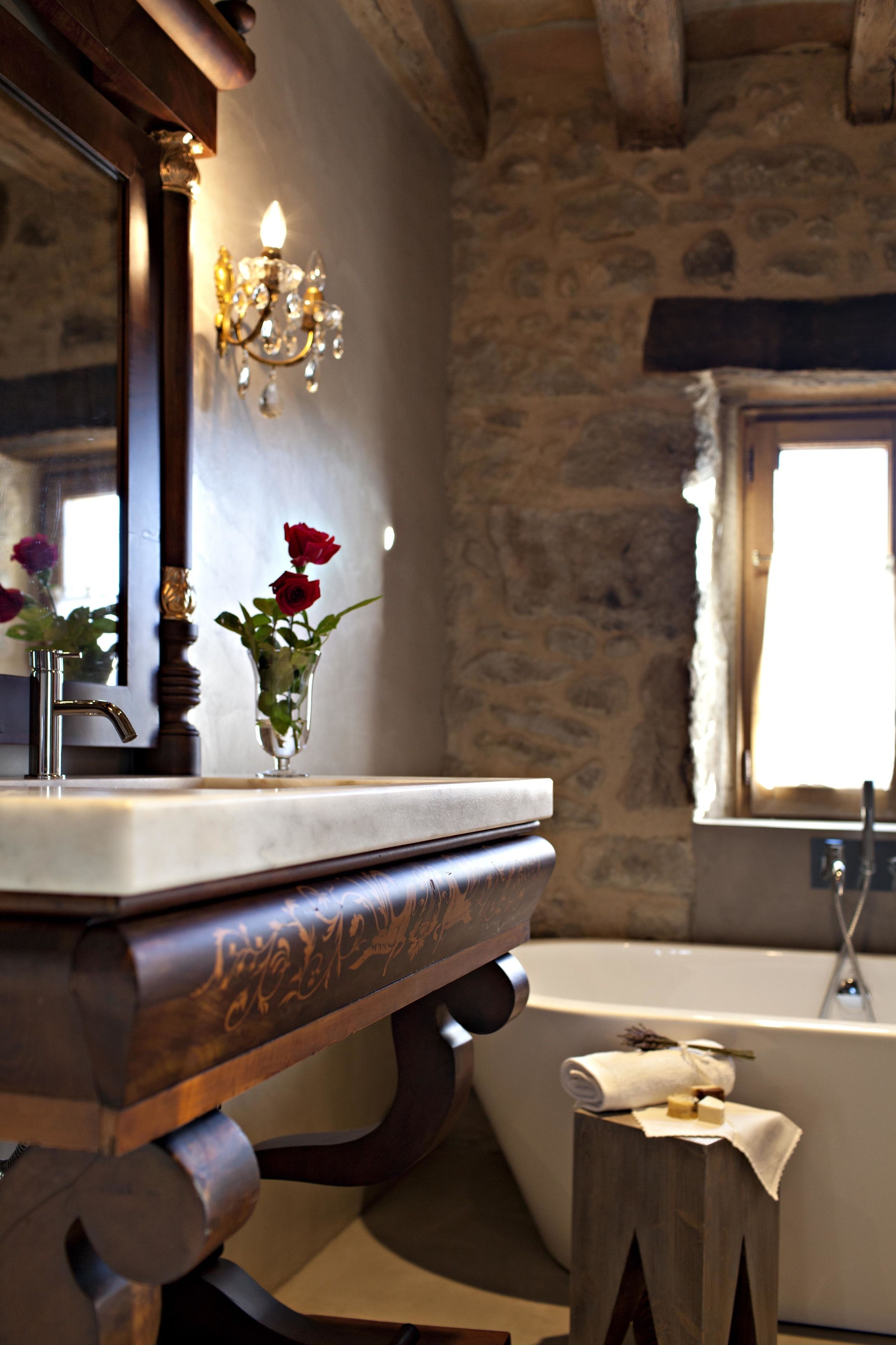 Un baño con pared de piedra, tocador de madera, espejo, candelabro y bañera.