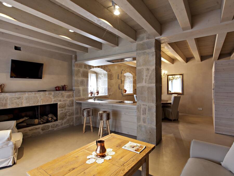 Un intérieur rustique avec des murs en pierre apparentes, une cheminée, une table en bois ornée d’un vase et un téléviseur fixé au mur.