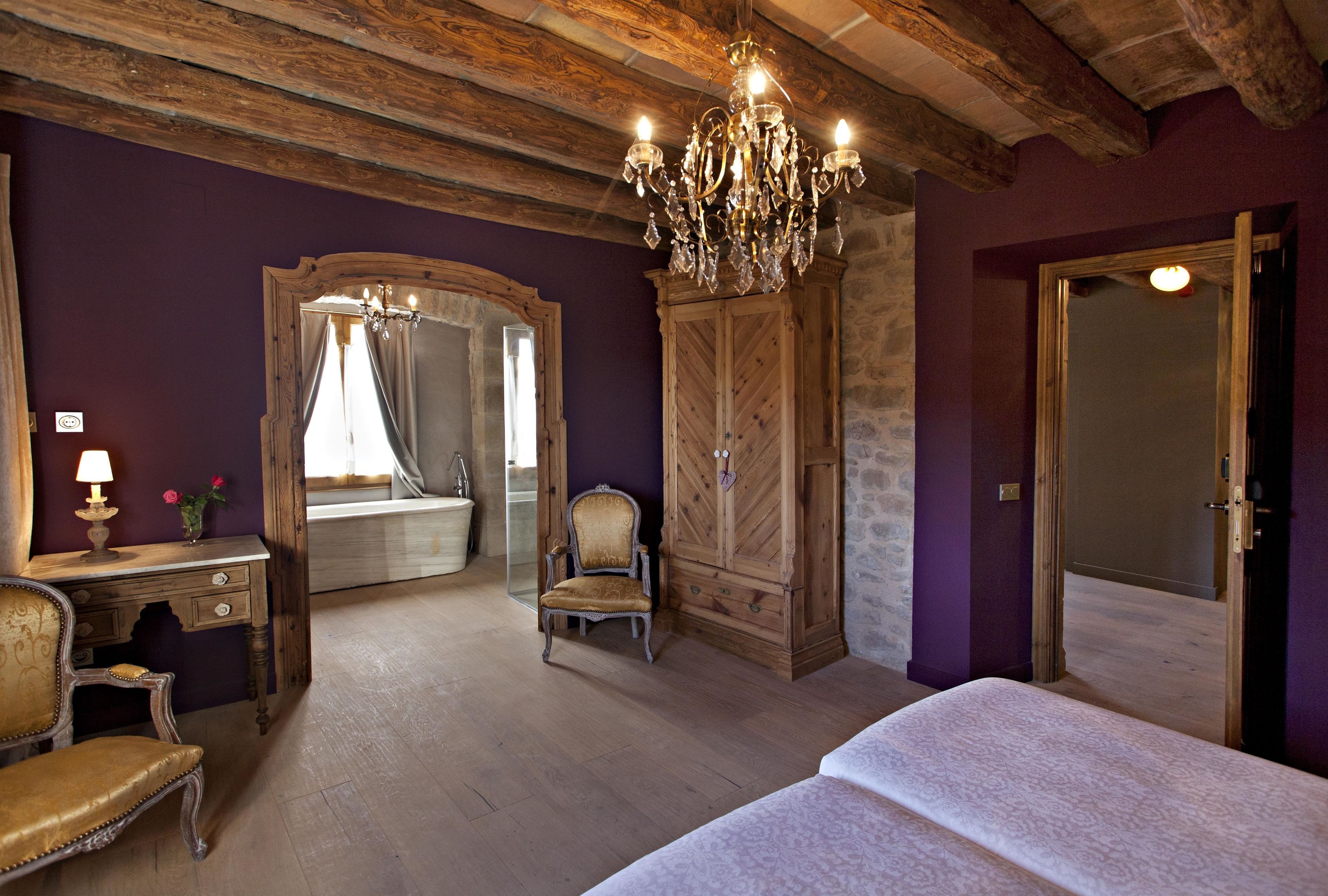 A bedroom with a wooden ceiling, a chandelier, a bed, a dresser, a chair, and a mirror.