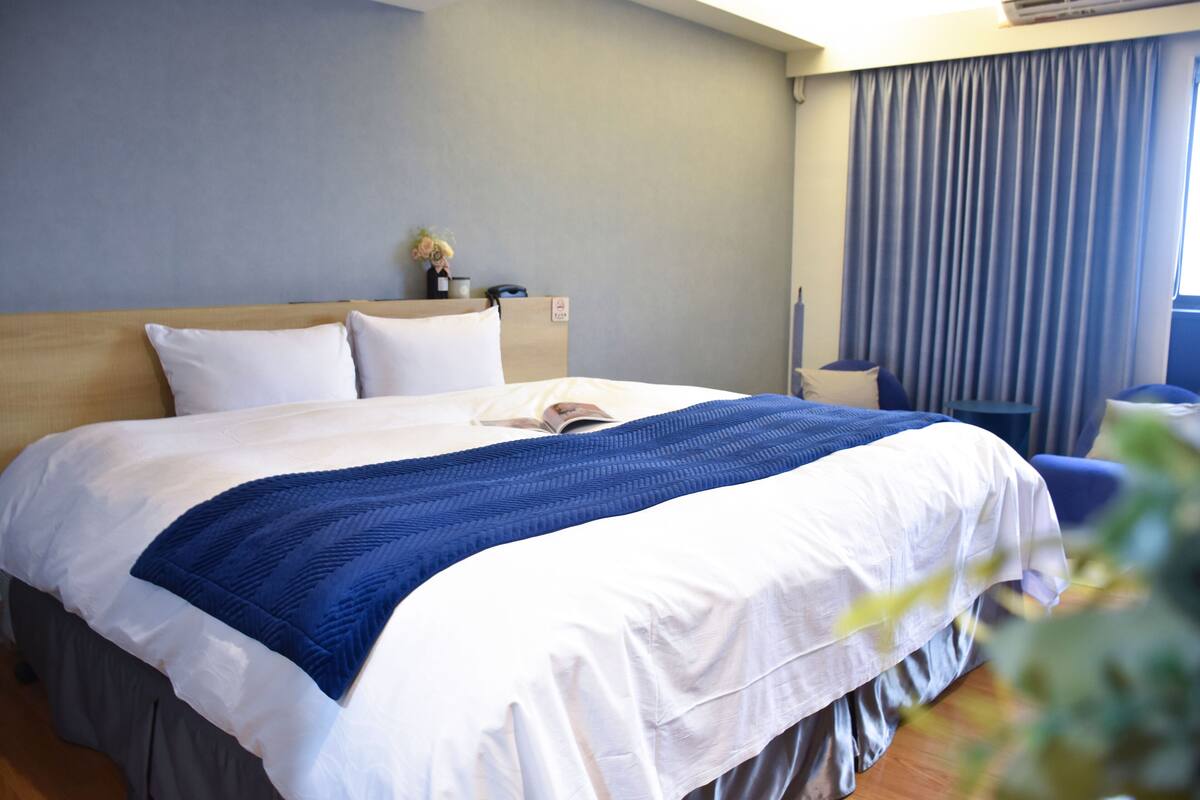 Quarto de hotel com uma cama grande, mesa de cabeceira, abajur e janela com cortinas.