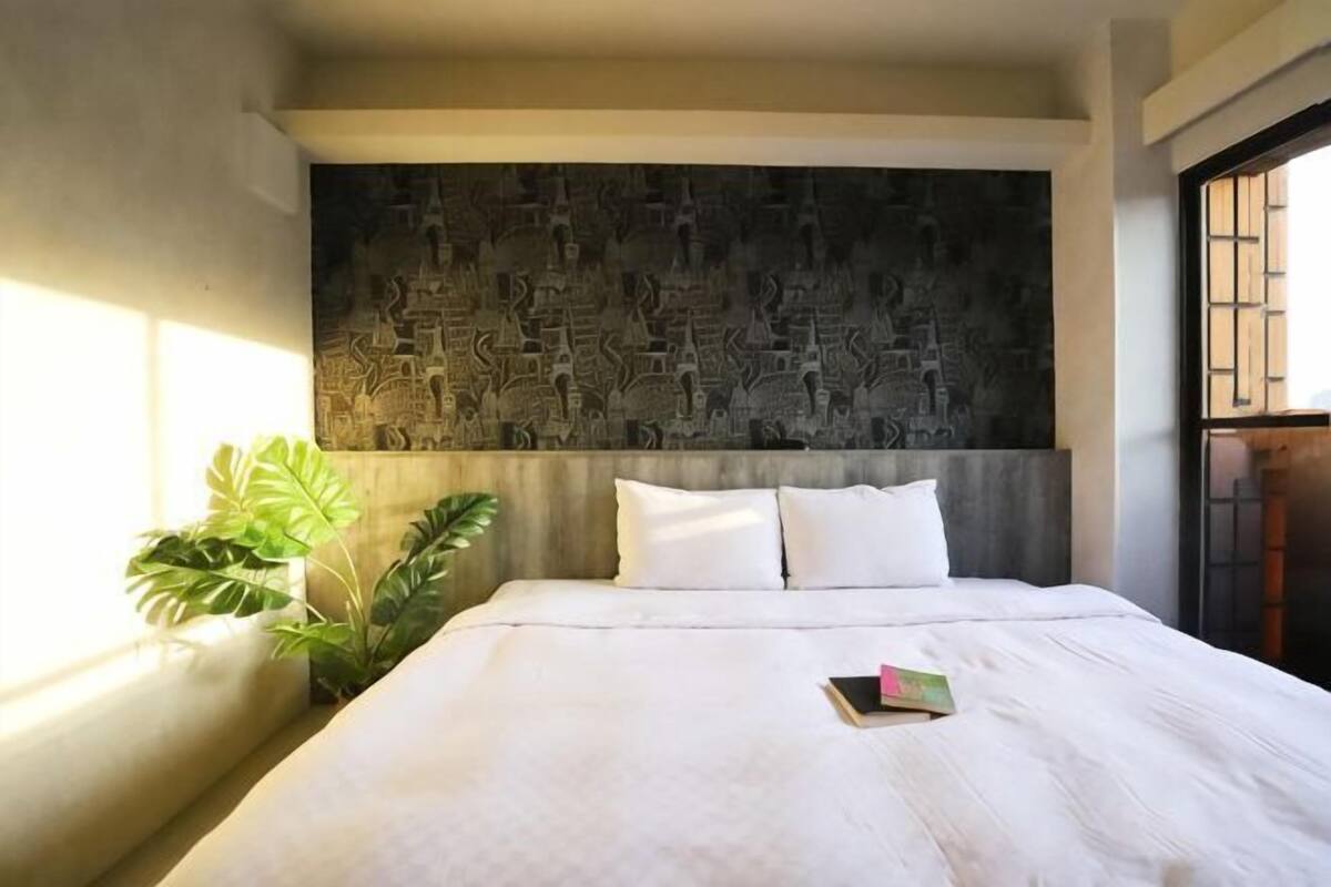 Uma cama com roupa de cama branca, cabeceira com design texturizado e um vaso de planta ao lado da janela.