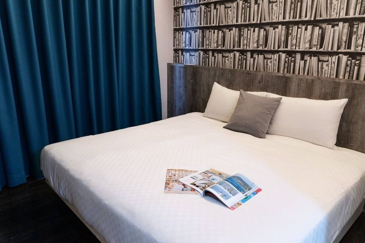 Cama bem arrumada com uma revista, uma estante de livros ao fundo e uma cortina azul à esquerda.