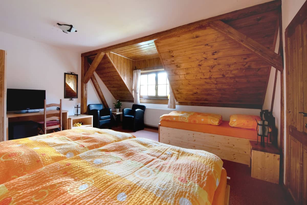 Un dormitorio con techo inclinado, vigas de madera, una cama con colcha estampada en naranja y amarillo, un escritorio con computadora, una silla y un televisor.