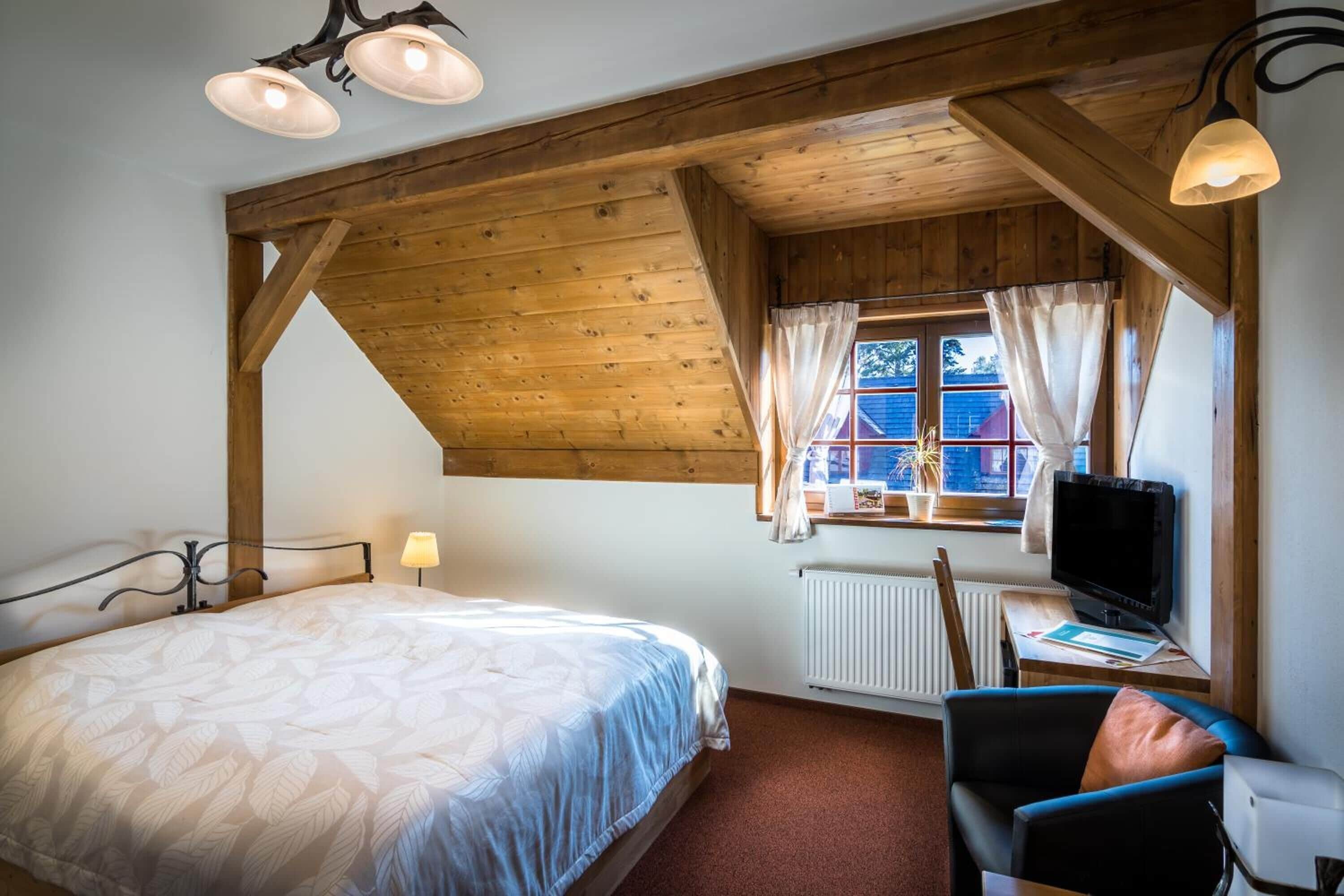 Quarto com teto de madeira, cama com colcha estampada com folhas, escrivaninha com computador e janela com cortinas.