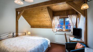 Quarto com teto de madeira, cama com colcha estampada com folhas, escrivaninha com computador e janela com cortinas.