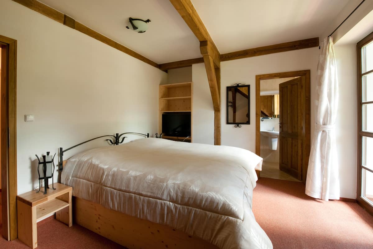 Un dormitorio con cama, cabecero de madera, una pequeña banca de madera, televisión, espejo y ventana con cortinas.