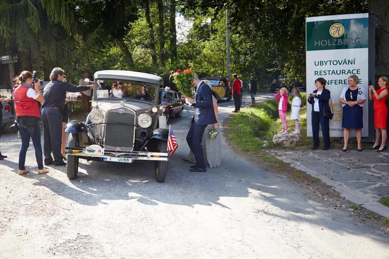 A vintage car parked on a road with people gathered around, a sign for 'Holzberg Ubytovany Restaurant Conference,' and a natural, tree-lined setting.