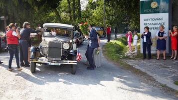 A vintage car parked on a road with people gathered around, a sign for 'Holzberg Ubytovany Restaurant Conference,' and a natural, tree-lined setting.