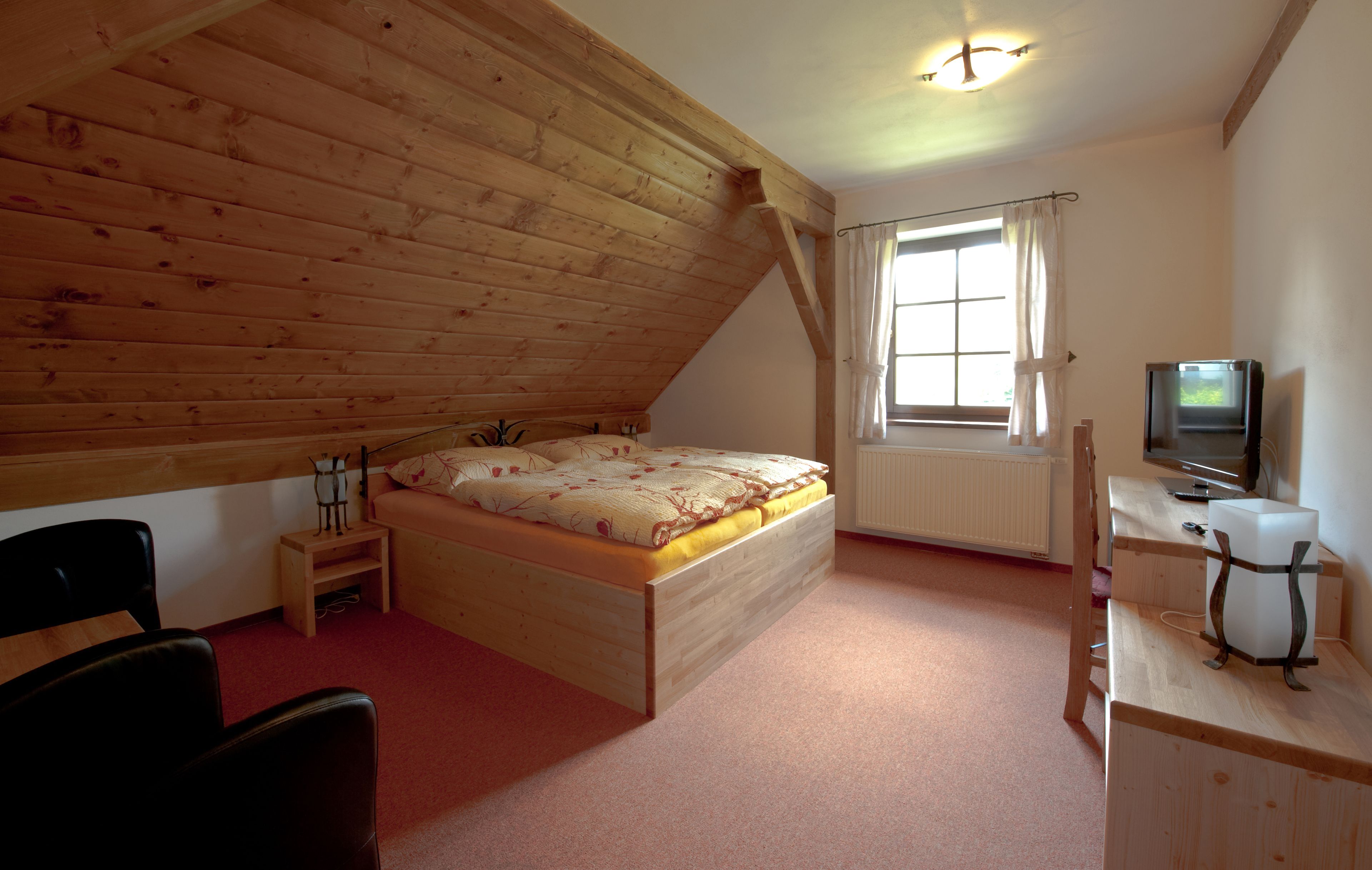 A bedroom with a sloped ceiling, wooden bed, desk, chair, and a television.