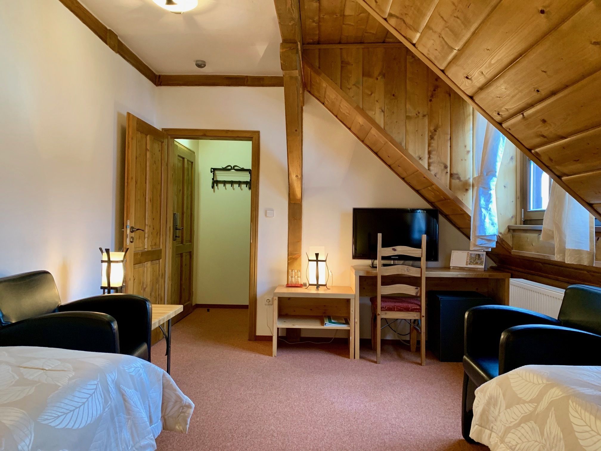 Una habitación acogedora con techo de madera, un televisor sobre un mueble de madera, un escritorio con una silla y dos camas con cubrecamas blancos.
